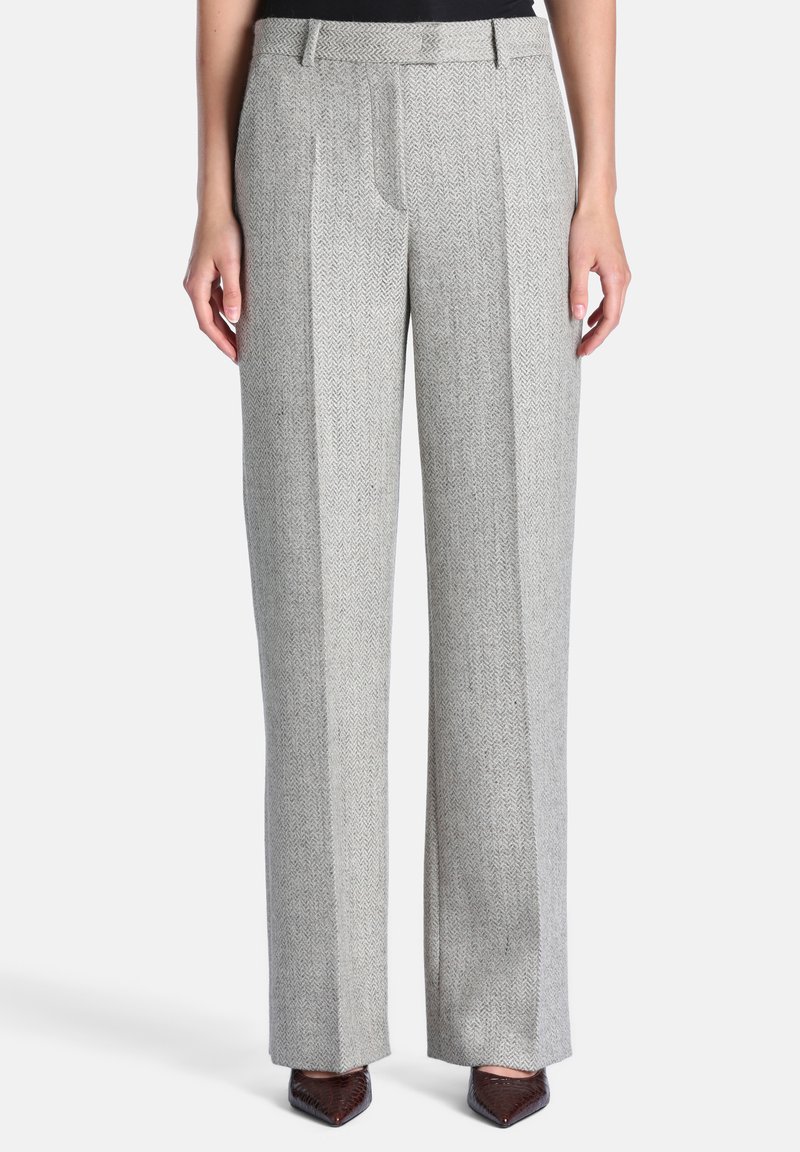 Pantalon gris clair à jambes larges avec un motif subtil en chevrons, porté avec des chaussures marron à bouts pointus et un haut noir visible à la taille.