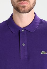 Camisa polo morada con tejido texturizado, tapeta con tres botones y un pequeño logo bordado de cocodrilo en el lado izquierdo del pecho.