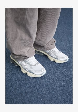 Valkoiset verkkomateriaalista valmistetut lenkkarit hopeisilla yksityiskohdilla, joissa on paksu pohja ja nauhoitusjärjestelmä. Yhdistetty harmaiden verryttelyhousujen kanssa. Sininen maton tausta.