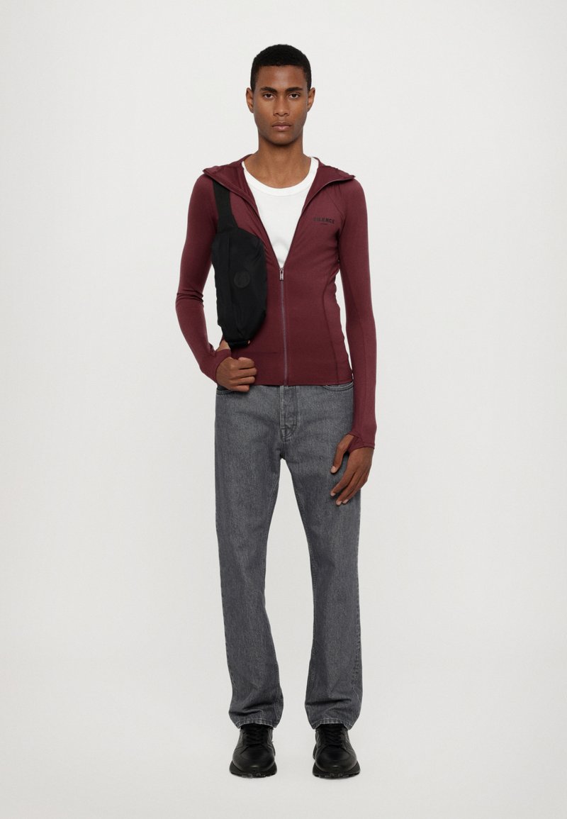 Bordo zip-up dukserica s dugim rukavima, uparena s sivim trapericama. Slim-fit dizajn, nošena s crnom torbom preko tijela i crnim cipelama.