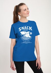 Camiseta de algodón azul con un gráfico de tiburón y texto blanco. El diseño incluye elementos náuticos y la frase "SNACK ATTACK". Mangas cortas.