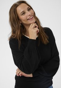 Femme aux longs cheveux bruns sourit en posant son menton sur sa main, portant un pull noir à manches bouffantes et des boucles d'oreilles en or.