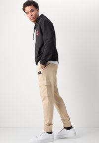 Svart huvtröja med logotyp, beige cargobyxor med fickor och vita sneakers. Mjukt material, avslappnad passform, ribbade muddar. Enkel färgpalett.