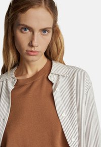 Witte shirt met fijne paarse strepen over een bruin gebreid sweater. Het shirt heeft een knoopsluiting aan de voorkant en een kraag, met een relaxte pasvorm.