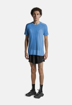 Jonge man staat met gezicht naar voren, draagt een blauw sport T-shirt, zwarte hardloopshorts, zwarte sokken en zwarte trailrunning schoenen tegen een witte achtergrond.