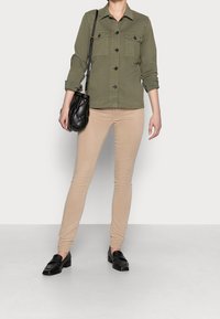 Olivengrøn button-down jakke med to brystlommer, beige skinny bukser og sorte loafers; bærer en sort quiltet håndtaske.