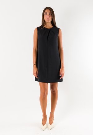 Donna in piedi su sfondo bianco che indossa un vestito nero senza maniche fino al ginocchio e décolleté bianche con punta a punta.