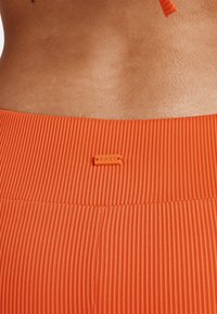 Narancssárga bordázott anyagú derékpánt, amelyen egy kis "ROXY" címke található. A textura sima, függőleges vonalakkal.