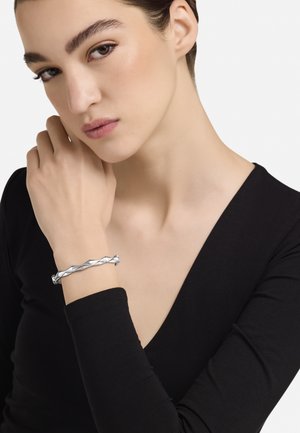 Srebrna uvrnuta narukvica glatke teksture, s jedinstvenim dizajnom s preklapajućim elementima, nošena na ruci uz neutralnu pozadinu.