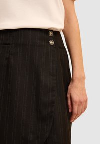 Zwarte pinstripe rok met twee decoratieve knopen in de taille, een getailleerde pasvorm en een gladde textuur. Hand rustend aan de zijkant.