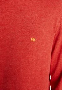 Pull en maille rouge avec un petit logo de machine à coudre jaune sur la poitrine gauche. Le tissu a une texture douce avec un léger motif chiné.