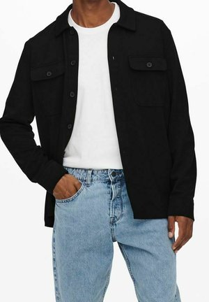 Uomo che indossa una giacca nera con bottoni sopra una t-shirt bianca e jeans a vita alta azzurri, con una mano in tasca, su sfondo bianco uniforme.