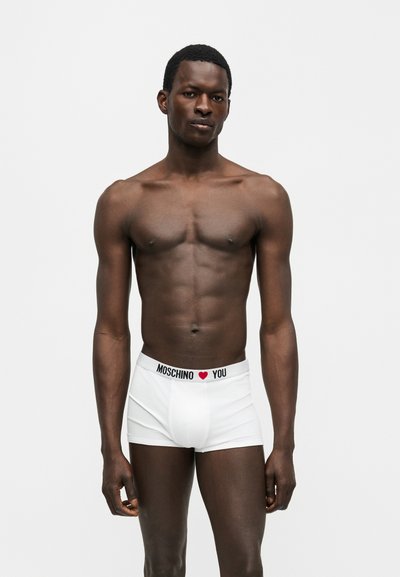 Slank mørkhudet mand står mod hvid baggrund iført hvide Moschino boxershorts med logo og rødt hjerte på linningen.