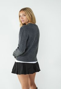 Grauer Strickpullover mit langen Ärmeln, rundem Ausschnitt und weicher Textur, kombiniert mit einem schwarzen Faltenrock. Einfaches Design, minimale Details.