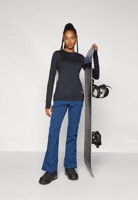 Black long-sleeve shirt with a round neckline, paired with blue snow pants and black snow boots. A snowboard is held next to the figure.