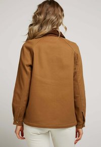 Veste marron texturée avec un col en velours côtelé, des poignets boutonnés et des manches raglan, présentée de dos sur un fond neutre.