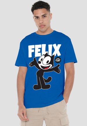 Young man wearing a blue T-shirt with a large Felix the Cat graphic and beige cargo shorts, standing with one hand in pocket.