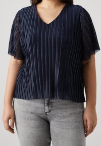 Donkerblauwe plooitop met korte, doorzichtige mouwen, V-hals en gestructureerde stof. Gemaakt met lichtgrijze skinny jeans.