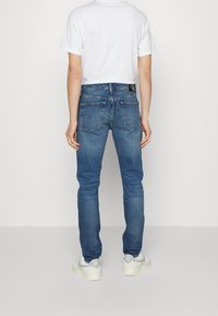 Person som står med ryggen mot kameran, klädd i en vit T-shirt, blå jeans med bakfickor och vita Nike Air-sneakers mot en enfärgad bakgrund.