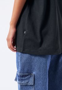 Černé bavlněné tričko se rovnými střihy, na boku s malou štítkovou logovací nášivkou. V kombinaci s volnými, světle modrými denimovými cargo kalhotami se bočními kapsami.