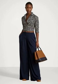 Γυναίκα που φοράει μια μοτίβο μπλούζα με μακριά μανίκια, ανοιχτό παντελόνι ναυτικού, καφέ ζώνη, καστανά παπούτσια και κρατάει μια καφέ και μαύρη τσάντα.