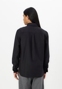 Camicia nera a manica lunga con colletto a bottoni, cucitura sulla spalla posteriore e maniche con polsini. Tessuto morbido, design aderente, abbinata a pantaloni a quadri grigi.