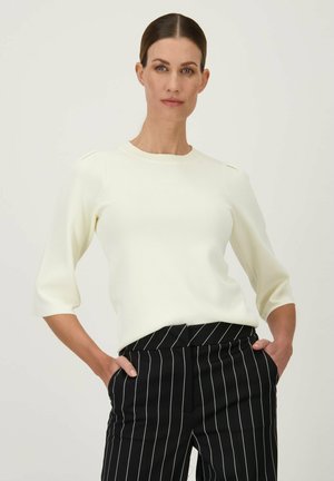 Pull blanc à col rond avec des manches trois-quarts et un détail aux épaules, associé à un pantalon à rayures noires et blanches. texture lisse et ajustée.