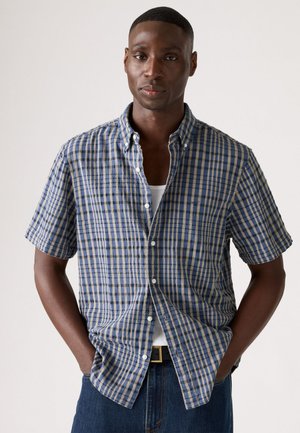 Camisa de cuadros de manga corta en azul y beige, hecha de tela suave, con cuello abotonado y bolsillo en el pecho, llevada sobre una camiseta sin mangas blanca.