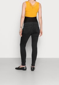 Žena otočená zády, oblečená v hořčicovém bezrukávovém topu s krajkovým zadním dílem, černých vysokých džínách, černých plochých botách a viditelnými tetováními na pažích a horní části zad.