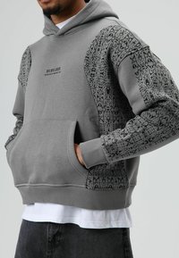 Grijze capuchontrui met zwarte printaccenten op de mouwen, een buidelzak aan de voorkant en de tekst "BYE BYE LOSER" op de borst. Zachte textuur.