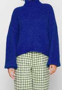 Strickpullover mit Rollkragen in lebhaftem Blau, mit weiten Ärmeln und lockerer Passform. Kombiniert mit Hosen im grün-weißen Hahnentritt-Muster.