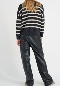 En blond person som har på sig en svart och beige randig tröja och svarta läderbyxor, stående mot en enkel vit bakgrund.