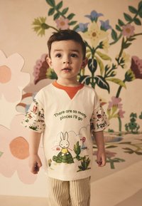 Balti marškinėliai su oranžiniu kaklu, vaizduojantys animacinius zuikio ir kaktusų piešinius bei tekstą. Trumpomis rankovėmis pagražinta spalvotais raštais, derinama su dryžuotomis kelnėmis.