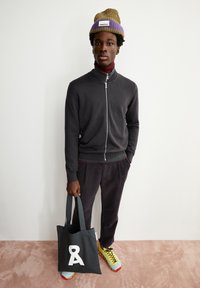 Schwarzer Reißverschluss-Pullover, bordeauxfarbener Rollkragenpullover darunter, graue Kord-Hosen, gelbe und rote Turnschuhe und eine grün-lila Mütze. Hält eine schwarze Tragetasche.