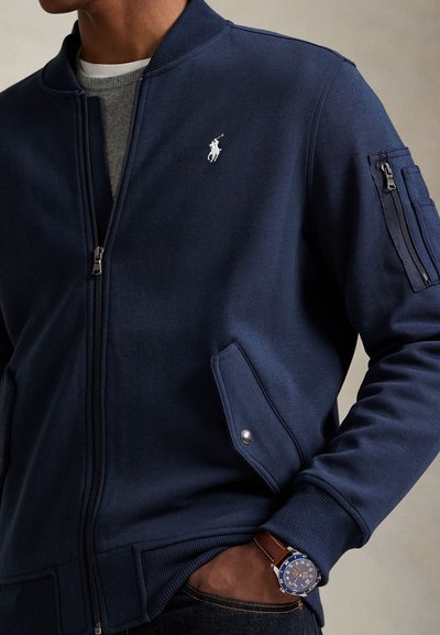 Homme portant une veste zippée bleu marine avec poche et logo polo blanc, chemise grise en dessous et montre-bracelet avec bracelet en cuir marron.