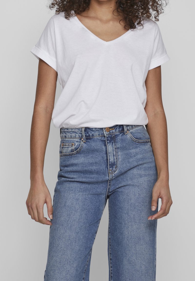 T-shirt blanc en V fabriqué en tissu doux, associé à un jean en denim bleu taille haute avec un design classique à cinq poches et une fermeture à bouton.
