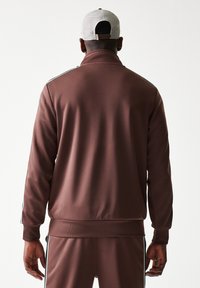 Καφέ αθλητικό μπουφάν με ψηλό γιακά, υφή και αντίθετες ρίγες στα πλάγια. Συνδυασμένο με παντελόνι στον ίδιο τόνο και γκρι καπέλο.