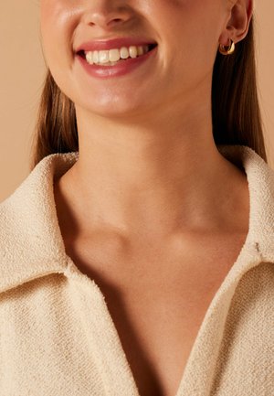 Femme souriante portant un haut texturé crème à col et de petites boucles d'oreilles anneaux dorés sur un fond beige.