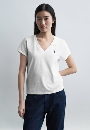Donna con capelli lunghi e scuri che indossa una T-shirt bianca con scollo a V con un piccolo logo ricamato blu e jeans blu scuro, in piedi davanti a uno sfondo semplice.