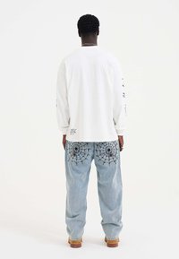 Langärmliges weißes Hemd mit schwarzem Text auf den Ärmeln; hellblaue Jeans mit Spinnennetz-Stickerei auf den Gesäßtaschen; hellbraune Schuhe.