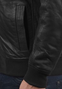 Musthave nahkajope, mille varruka ja soonikko on tumedast nahast, käsi on siniste teksade tasku lähedal.