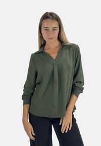 Donkergroene blouse met lange mouwen en kraag, voorzien van een geplooide voorkant en een zachte textuur, gecombineerd met een zwarte broek.
