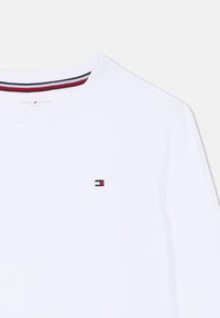 Hvid langærmet skjorte med rund halsudskæring, prydet med et lille broderet logo i rød, hvid og blå på brystet.