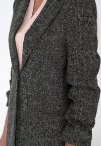 Teksturowany szary żakiet z wyciętym kołnierzem, wyposażony w jedną przednią kieszeń i lekko marszczone rękawy, noszony na jasnej bluzce.