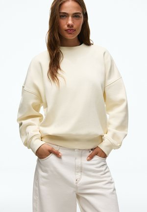 Junge Frau mit hellbraunen Haaren trägt ein cremefarbenes Oversized-Sweatshirt und weiße hoch taillierte Hosen, die Hände in den Taschen.