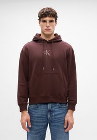 Brun hettegenser i myk stoff med kengurulomme og snøring. Inkluderer en liten logo plassert midt på brystet. Casual passform.