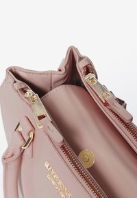 Lichtroze handtas met een gestructureerde buitenkant, twee gouden ritsen en gouden hardware-accenten. Heeft een ronde vorm en een ruime binnenkant.