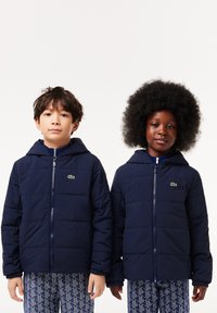 Vestes matelassées bleu marine avec capuches, dotées de fermetures éclair et de poignets élastiques. Logo Lacoste sur la poitrine gauche. Pantalons à motif géométrique texturé.