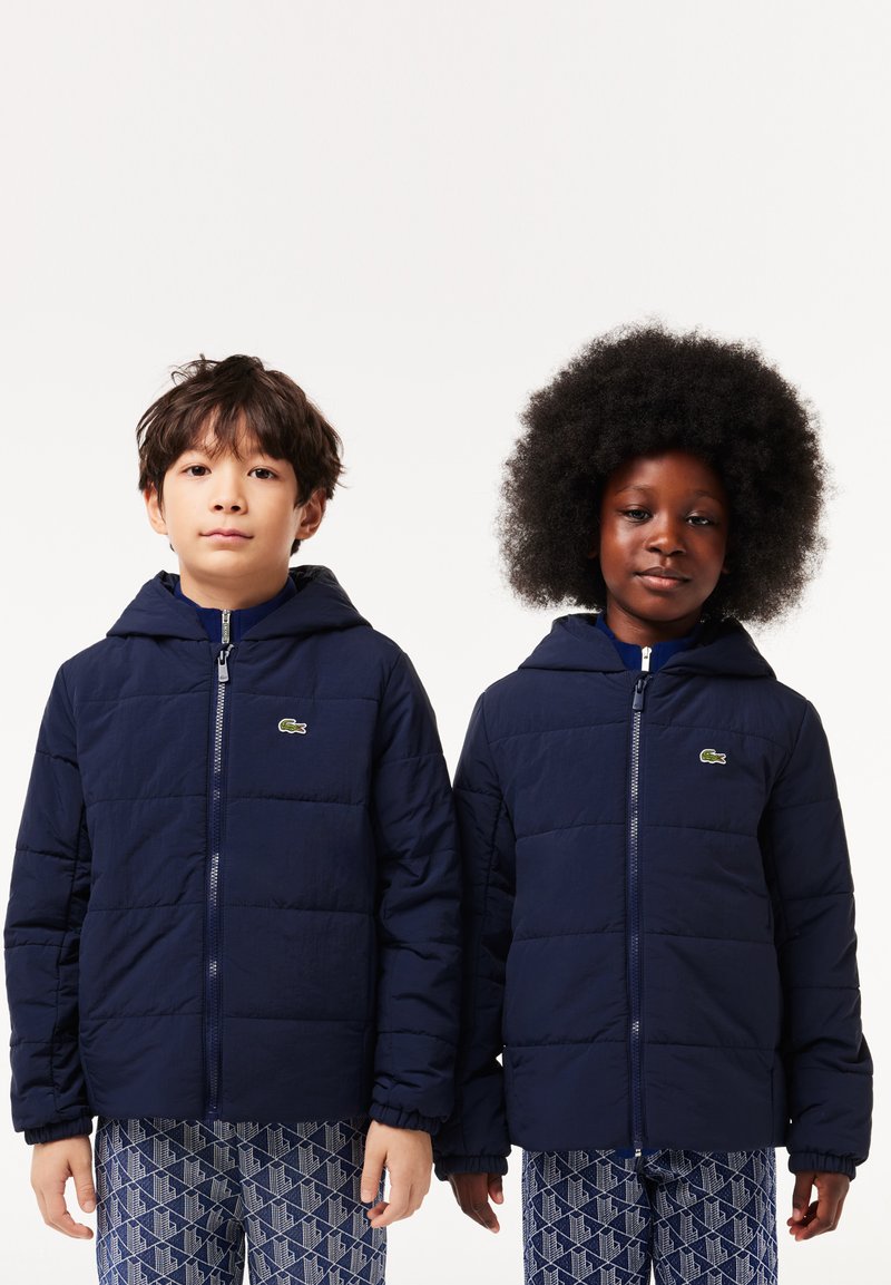 Marineblaue gepolsterte Jacken mit Kapuzen, ausgestattet mit Reißverschluss und elastischen Bündchen. Lacoste-Logo auf der linken Brust. Texturierte geometrisch gemusterte Hosen.