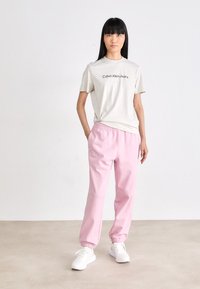 Svetlo béžové tričko s nápisom "Calvin Klein Jeans" vytlačeným čiernou farbou, skombinované so svetloružovými teplákmi s elastickými manžetami a postrannými vreckami. Biele tenisky.
